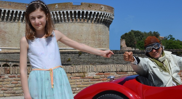 Elena Schiavoni nei panni della bambina, protagonista del ritrovamento delle bobine con le corse, mentre saluta il passaggio di un auto storica davanti alla Rocca roveresca di Senigallia